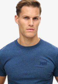Blauwe katoenen T-shirt met ronde hals; heeft een subtiele heathertextuur en een klein geborduurd logo op de borst.