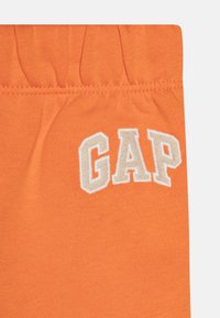 Oranžové látkové šortky s elastickým pásom, na ktorých je vyšitý logo "GAP" béžovou farbou. Hladká textúra a jednoduchý dizajn.
