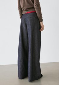 Pantalon en denim foncé à jambes larges avec une texture lisse, doté d'une poche arrière et d'une ceinture régulière visible au-dessus d'un haut rouge.