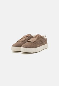 Baskets en suede marron avec semelles en caoutchouc blanches, présentant un profil bas, un embout arrondi et des lacets ton sur ton. Marquage subtil sur le côté.