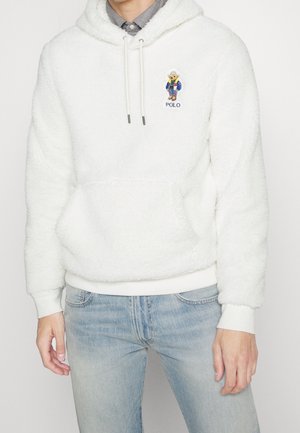 Hombre con sudadera blanca peluda con un pequeño oso bordado y texto "POLO", combinado con vaqueros azul claro y cuello de camisa a cuadros visible.