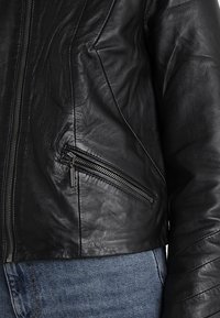 Černá kožená bunda s přiléhavým střihem, obsahující kapsu na zip a texturovaný materiál. Ukázáno s modrými džíny.