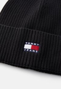 Crna pletena kapa s rebrastom teksturom. Ima mali komadić s natpisom "TOMMY JEANS" i obojene blokove bijele, crvene i plave boje.