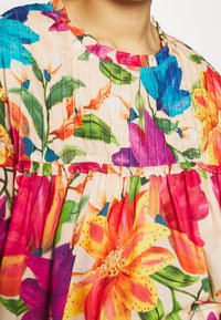 Bluse med blomsterprint, der har livlige flerfarvede blomster i pink, orange og blå på en lys baggrund, med plisseret tekstur og flæsekrave.