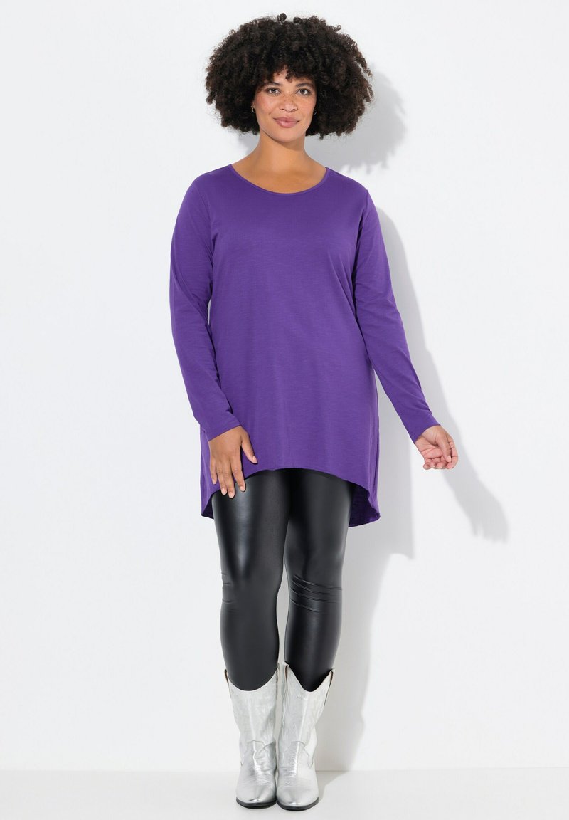 Lila Langarm-Tunika mit abgerundetem Saum, kombiniert mit schwarzen Leggings und silbernen westernstil Boots, vor einem weißen Hintergrund präsentiert.