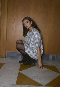 Haljina na crno-bijele kvadrate s kratkim rukavima, nosi se s crnim neprozirnim tajicama i tamnosmešnim čizmama s petom, u pozadini od drvenih panela.