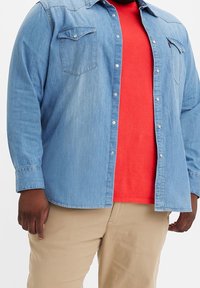 Mężczyzna w jasnoniebieskiej koszuli dżinsowej z guzikami nałożonej na czerwoną koszulkę oraz beżowe spodnie, stojący na tle białym.