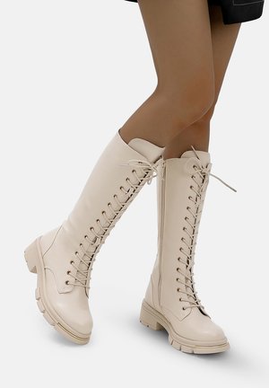 Bottes crème à lacets montant jusqu'au genou, fabriquées en cuir lisse, avec des œillets dorés, une semelle épaisse et une pointe arrondie.