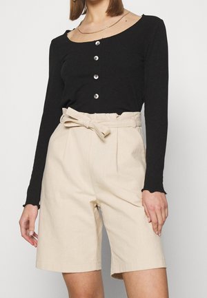 Vrouw met een zwart, lang mouwig shirt met knopen en beige shorts met hoge taille, voorgebonden met een stoffen ceintuur.