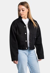 Chaqueta de mezclilla negra recortada con cuello puntiagudo, botones frontales y dos bolsillos en el pecho. Usada sobre una camisa blanca y unos jeans de color azul claro.