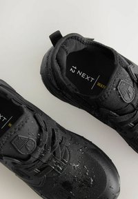 Chaussures noires imperméables avec une surface texturée, des lacets et des détails accentués. Gouttes d'eau visibles sur le matériau. Taille 12 étiquetée à l'intérieur.