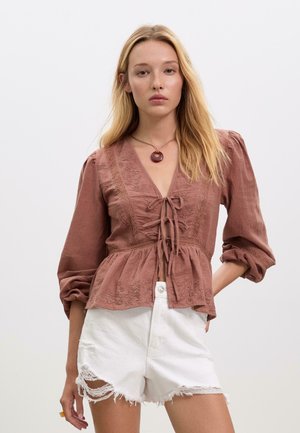 Blouse marron à col en v avec broderies florales, détails noués à l'avant, manches ballons. Assortie avec un short en denim blanc déchiré.