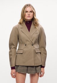 Blazer castanho de double breasted com lapelas notched, bolsos laterais e botões pretos. Usado sobre um suéter de gola alta em cor bordô e uma saia às riscas.