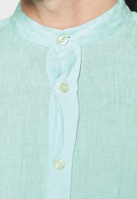 Camisetta verde chiaro a maniche corte in lino, con colletto alla mandarino e due bottoni frontali, caratterizzata da un sottile motivo tessile.