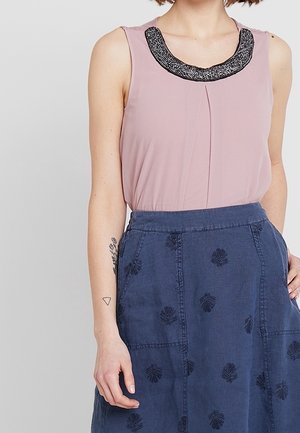 Mouwloze roze blouse met een zwarte kralenrand bij de hals, gecombineerd met een blauwe rok met donkere bladborduursels en twee voorzakken.
