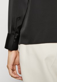 Svart silkesblus med en slät textur, med uppvikt ärm, två knappar och en nederkant som lätt draperar sig över ljusa byxor.
