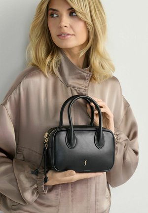 Blondynka w beżowej satynowej kurtce trzymająca małą czarną skórzaną torebkę z dwoma uchwytami i złotym suwakiem.