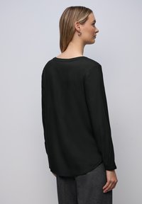 Blusa nera a maniche lunghe con scollo rotondo, texture liscia e lieve drappeggio sul retro. I dettagli includono un polsino discreto alle maniche.