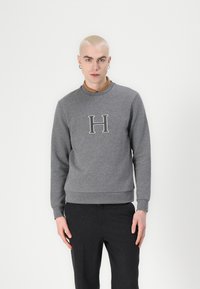 Grauer Pullover aus einem Baumwoll-Mix, mit einem runden Halsausschnitt und einem großen bestickten "H" auf der Vorderseite. Mit Bündchen an den Ärmeln und Saum.