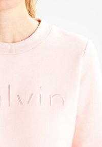 Sudadera de color rosa claro con un cuello acanalado. Presenta texto en relieve "calvin" en un color a juego, fabricada en un suave material de algodón.