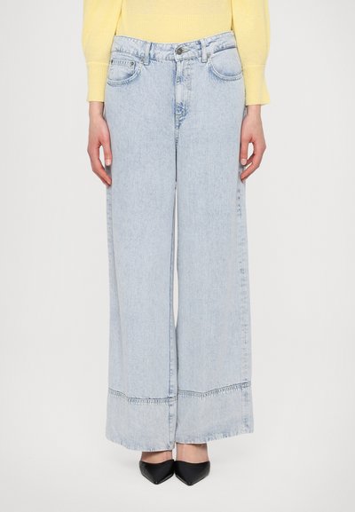 Jean large bleu clair en denim, avec une fermeture éclair, des poches avant et une couture visible le long de l'ourlet, assorti à des chaussures noires.
