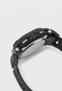 Reloj negro con una esfera redonda, correa de goma, herrajes plateados y tres botones a un lado. La parte posterior tiene un acabado metálico suave.