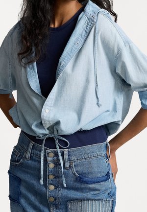 Kvinde iført en navyblå top under en lyseblå cropped jakke med snøre, parret med et højtaljet denimskørt med knapper og lapper.