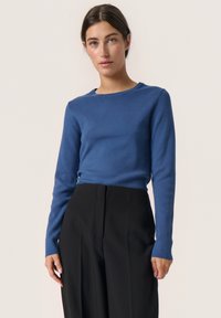 Blauer langärmliger Pullover in figurbetonter Passform mit Rundhalsausschnitt. Die Textur wirkt glatt und wird mit hochgeschnittenen schwarzen Hosen kombiniert.
