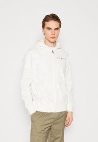 Felpa bianca con zip realizzata in tessuto morbido, con un logo frontale e un ampio cappuccio. Progettata con due tasche laterali e polsini a coste.