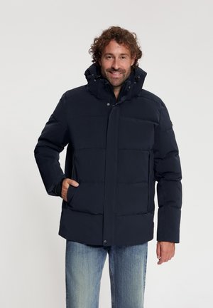 Man met krullend haar die een marineblauw gewatteerd jack draagt met handen in de zakken en lichtblauwe jeans, staand tegen een effen achtergrond.