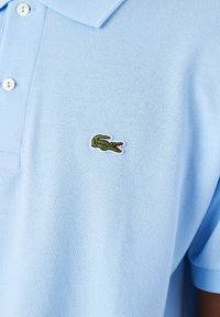 Polo shirt bleu clair en tissu texturé, avec un col structuré et un petit logo crocodile vert brodé sur la poitrine.