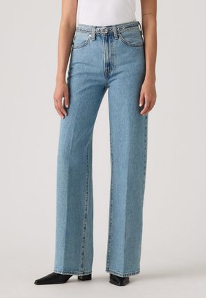 Person som har på seg høytlivs, lyseblå jeans med rette ben, belte og svarte sko med spiss tupp, stående mot en ensfarget bakgrunn.