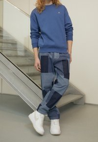 Modrá mikina s malým logem, džíny z různobarevných denimů v různých odstínech modré a bílé tenisky. Stojí na schodišti.