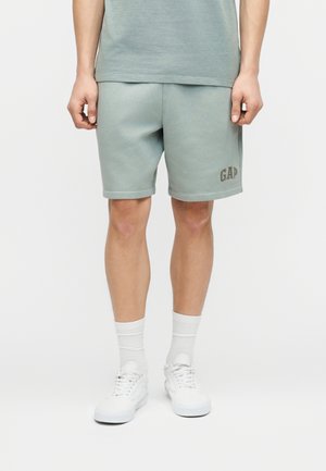 Hombre con pantalones cortos de color verde claro con el logo "GAP", calcetines blancos y zapatillas blancas, de pie contra un fondo blanco liso.