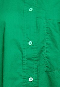 Camicia verde con bottoni, realizzata in tessuto liscio. Presenta bottoni di un verde chiaro e una tasca sul lato sinistro. La texture sembra leggermente stropicciata.
