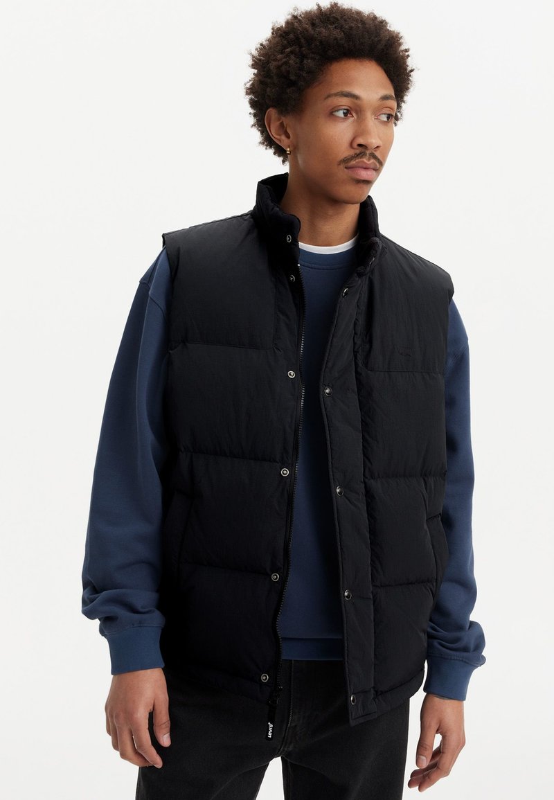 Svart puffer-väst med hög krage, knappstängning och quiltat mönster, bärs över en blå sweatshirt. Har sidodragkedja och minimalistisk design.
