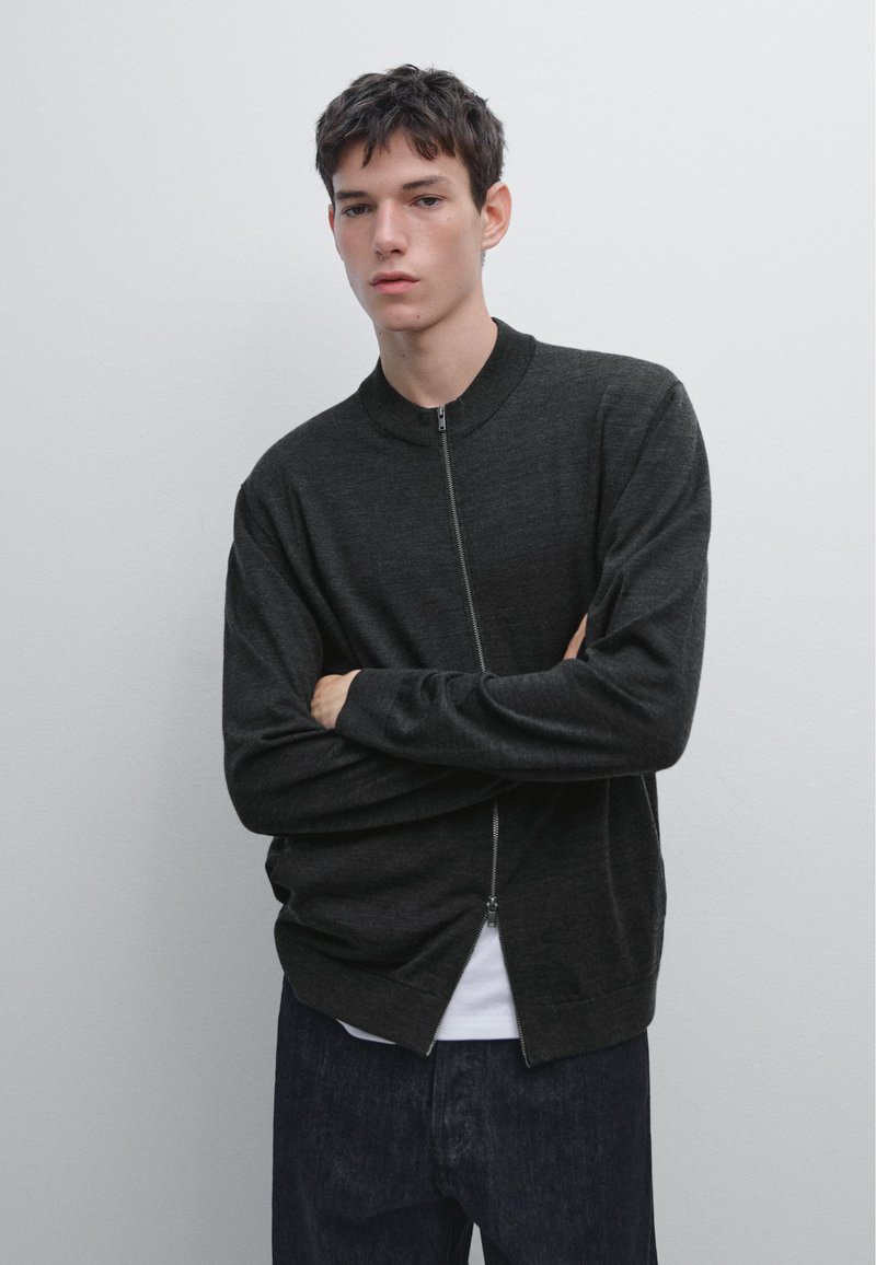 Cardigan zippé gris foncé avec un col rond, des manches longues et une coupe ample, porté par-dessus une chemise blanche et associé à un jean foncé.