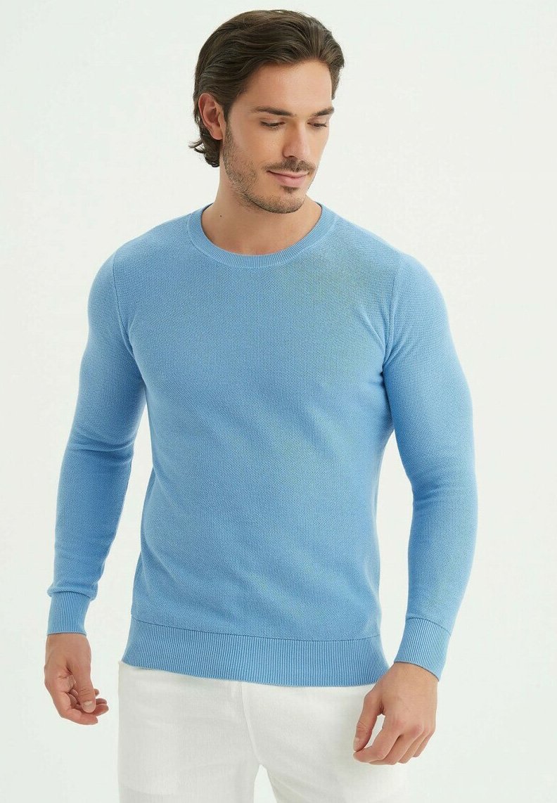 Pull en tricot bleu clair avec des manches longues et un col rond. Présente des poignets et un ourlet côtelés. Porté sur un pantalon blanc.