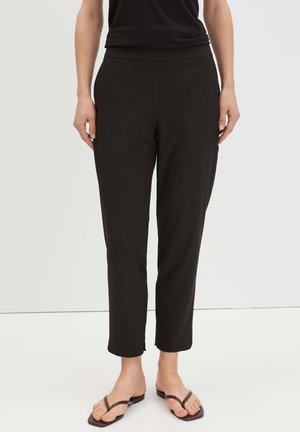 Vrouw draagt zwarte taps toelopende broek en bruine platte sandalen, staand tegen een effen witte achtergrond.