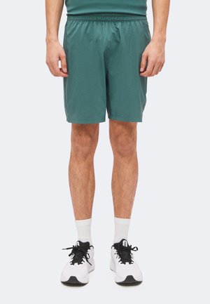 Homme portant un short de sport vert, des chaussettes blanches et des baskets blanches avec des lacets noirs, debout les bras détendus le long du corps.