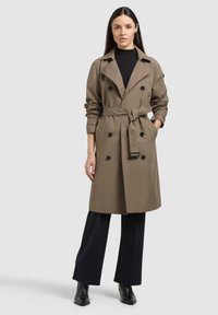 Donna con capelli lunghi e scuri che indossa un trench marrone con cintura, dolcevita nero, pantaloni neri a gamba larga e stivaletti neri su sfondo grigio.