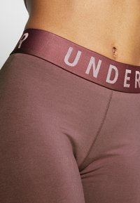 Brązowe legginsy z szerokim bordowym paskiem w talii z wytłoczonym napisem "UNDER". Tkanina jest gładka i dopasowana, z widocznymi detalami przeszyć.