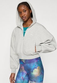 Γκρι cropped φούτερ με φερμουάρ και κουκούλα, με χαμηλούς ώμους και ελαστικά μανίκια, φορεμένο πάνω από ένα τιρκουάζ τοπ και πολύχρωμο κολάν με σχέδια.