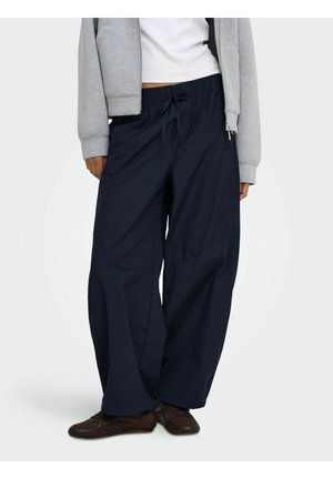 Personne portant un pantalon large bleu marine avec une taille à cordon, un haut blanc, une veste grise zippée et des chaussures marron foncé.