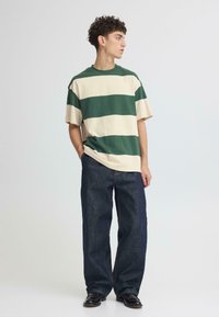 T-shirt en coton rayé vert et crème à manches courtes, associé à un jean large en denim foncé et des chaussures noires sur un fond neutre.