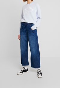 Ljusblå sweatshirt med texten "HOLLISTER", kombinerad med vida mörkblå denimjeans med fransar i nederkant och sidslitsar, bärs med svarta sneakers.