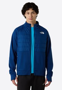 Modrá zipová bunda s texturovanou spodní částí a hladkými horními panely. Obsahuje vysoký límec, logo North Face a dlouhé rukávy.