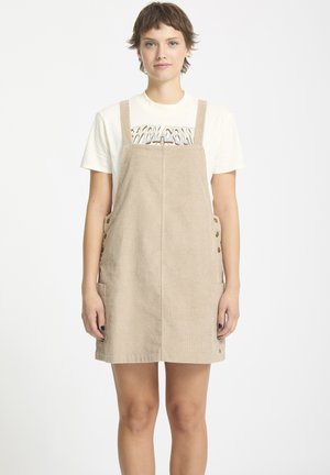 Beige corduroy tuinbroek jurk met verstelbare banden, voorzakken en knoopaccenten. Gedragen over een witte T-shirt met opdruk.