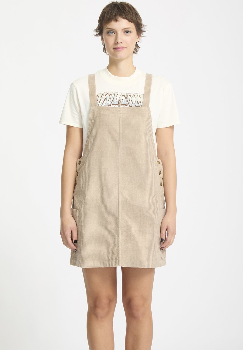 Beige corduroy tuinbroek jurk met verstelbare banden, voorzakken en knoopaccenten. Gedragen over een witte T-shirt met opdruk.