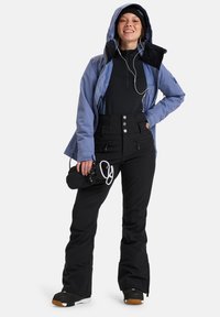 Lichtblauwe waterdichte jas met capuchon, zwarte langjarige top, hoge zwarte broek met knopen en houdt een klein accessoire vast.